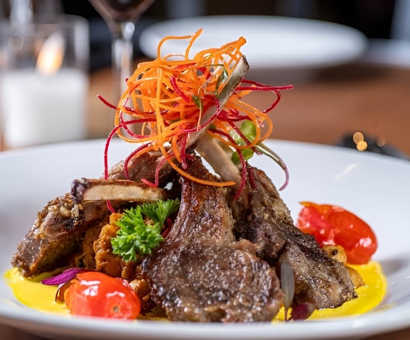 Grilled lamb chops with colorful shredded vegetables, cherry tomatoes, and yellow sauce on a white plate.