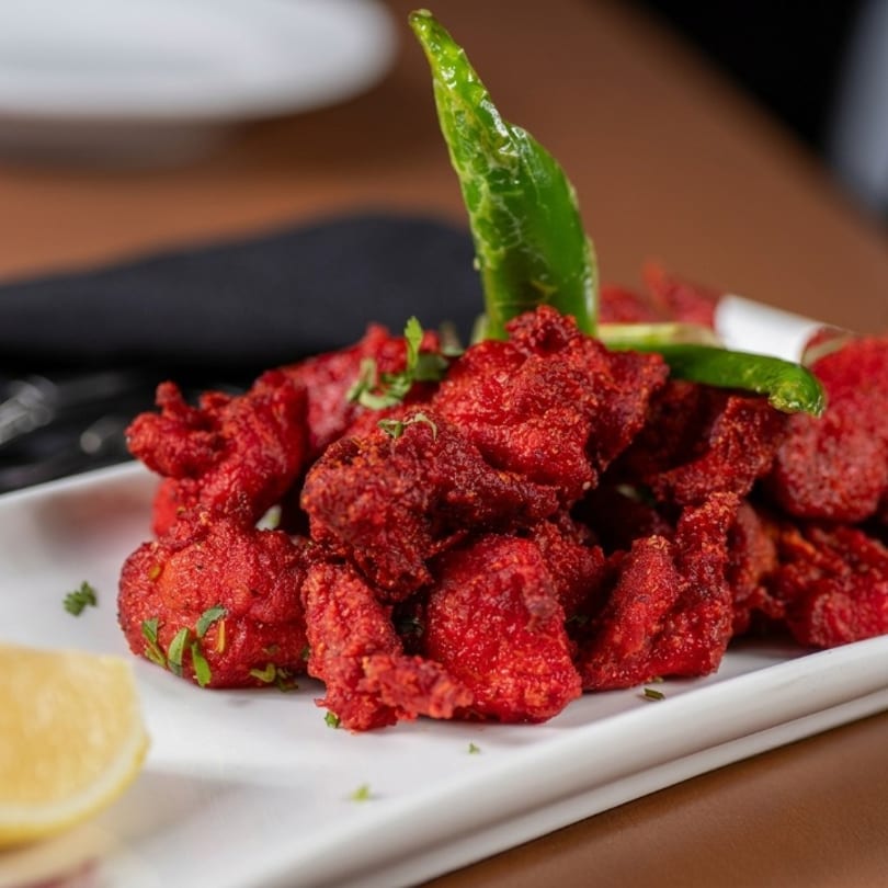 Red fried chicken pieces garnished with green chili, herbs, and a lemon wedge on a white plate.