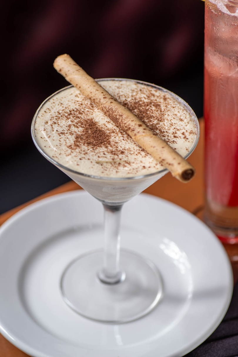 Creamy dessert in a martini glass, topped with cocoa powder and a wafer stick, on a white plate.
