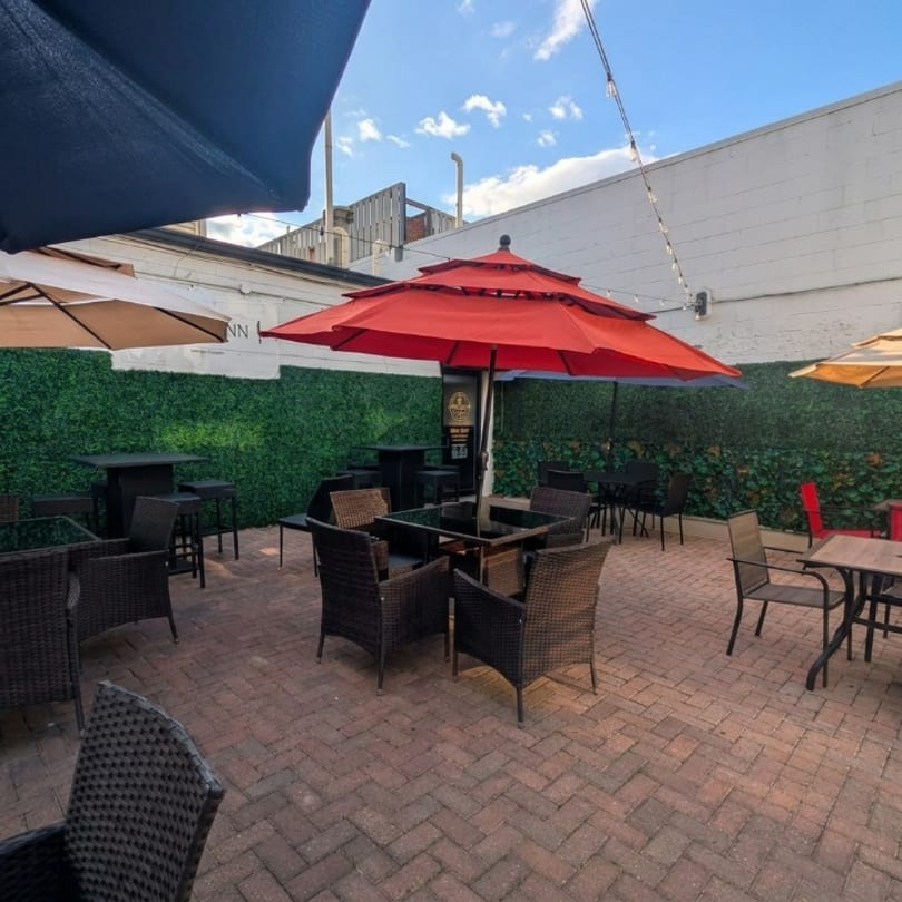 Outdoor restaurant patio with wicker chairs, tables, red and beige umbrellas, and a green hedge wall.