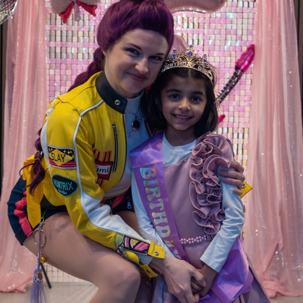 Smiling woman with purple hair and girl in a crown and birthday sash pose at a party.