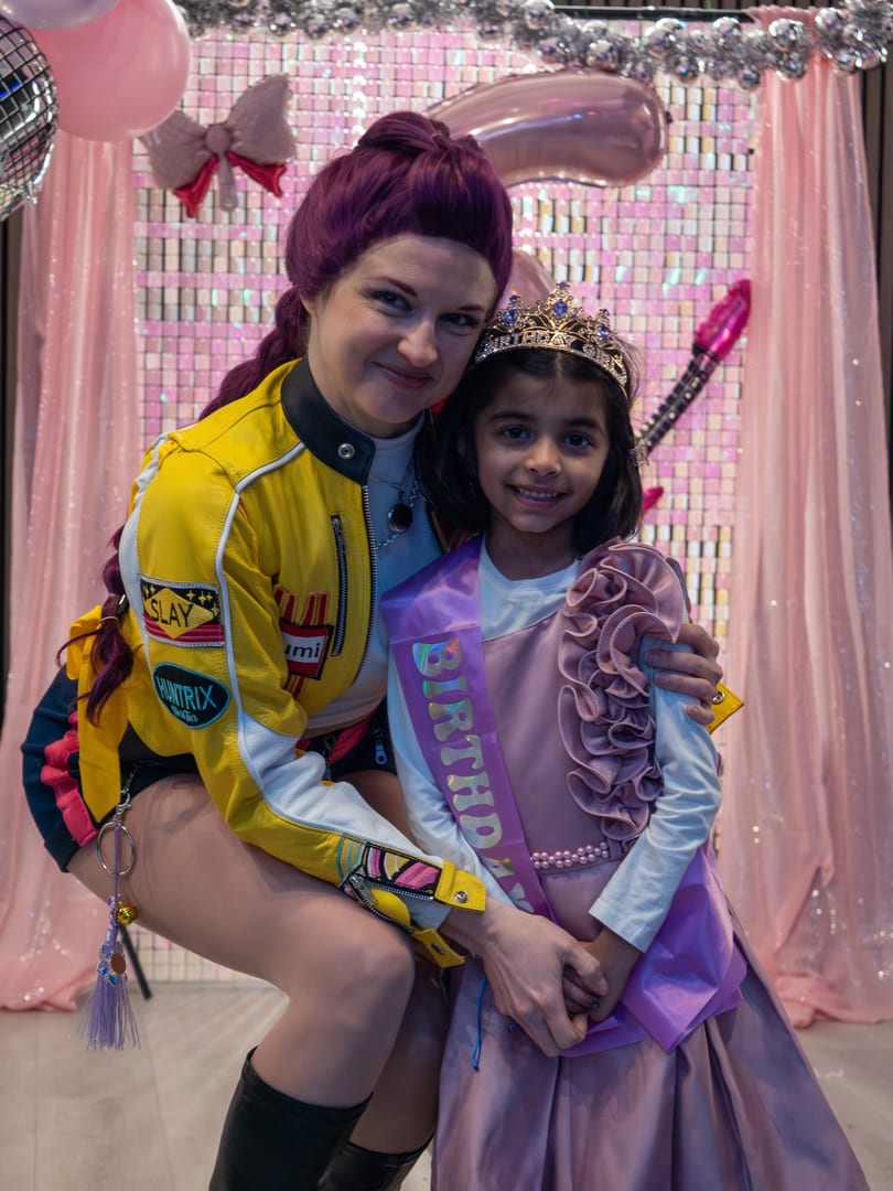 Smiling woman with purple hair and girl in a crown and birthday sash pose at a party.