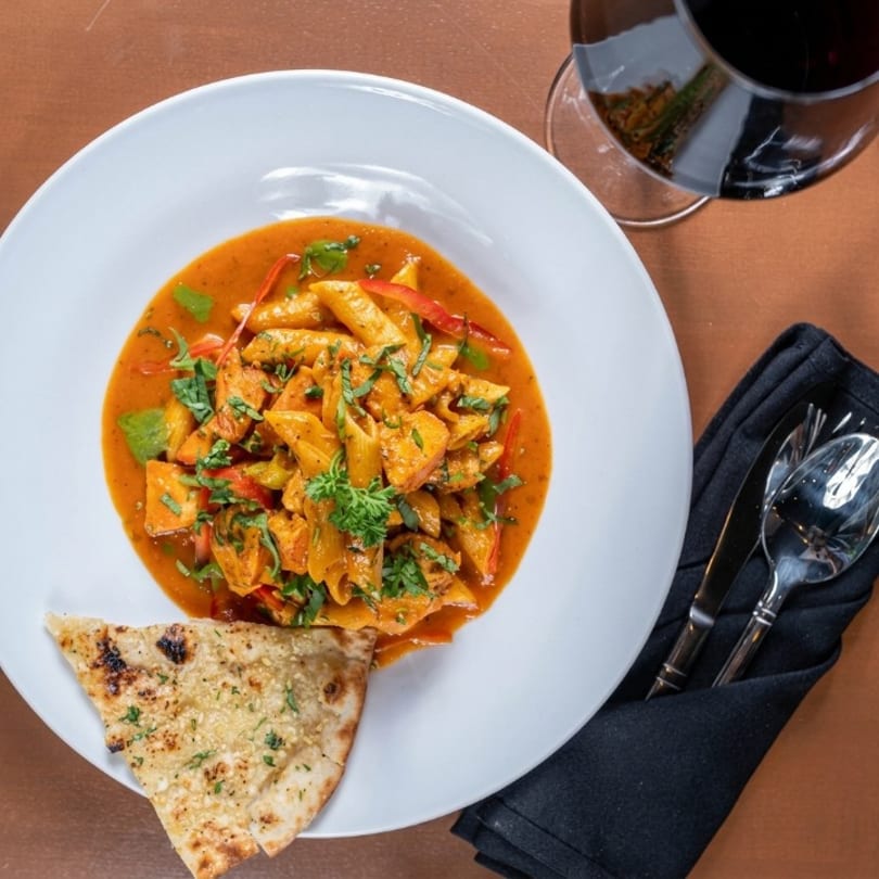 Penne pasta with chicken and red sauce, naan bread, red wine, and cutlery on a table.