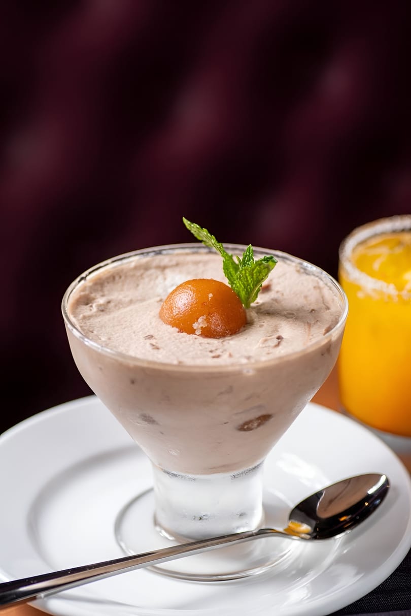 Chocolate mousse dessert with a gulab jamun and mint leaf, next to an orange drink.