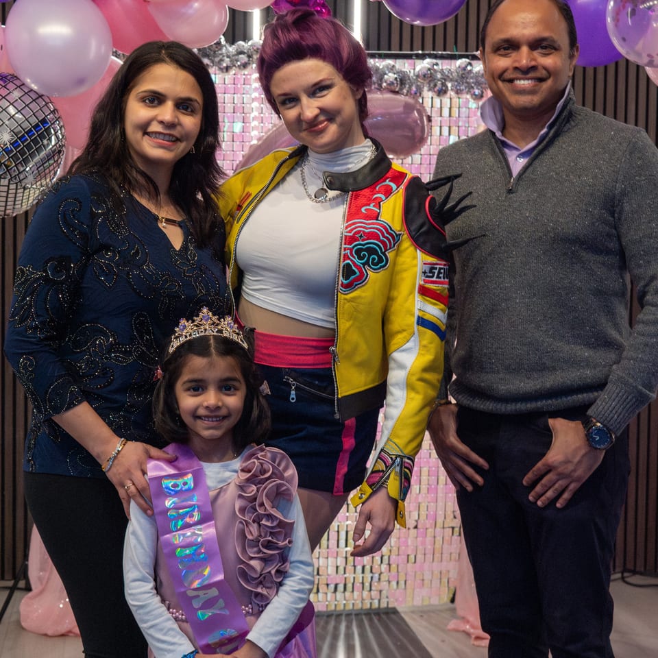 Two women, a man, and a young girl in a crown and birthday sash pose with balloons.