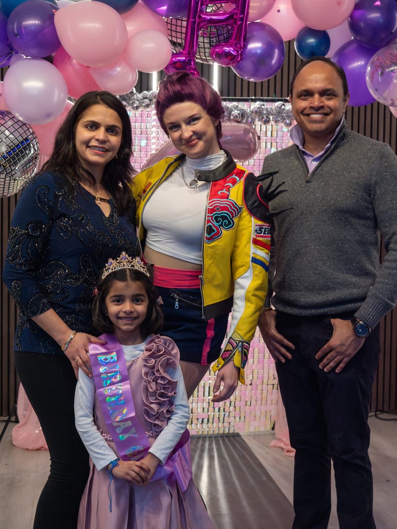 Two women, a man, and a young girl in a crown and birthday sash pose with balloons.