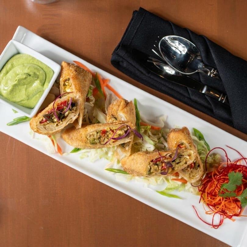 Crispy egg rolls with filling, green dipping sauce, shredded vegetables, and cutlery on a brown table.