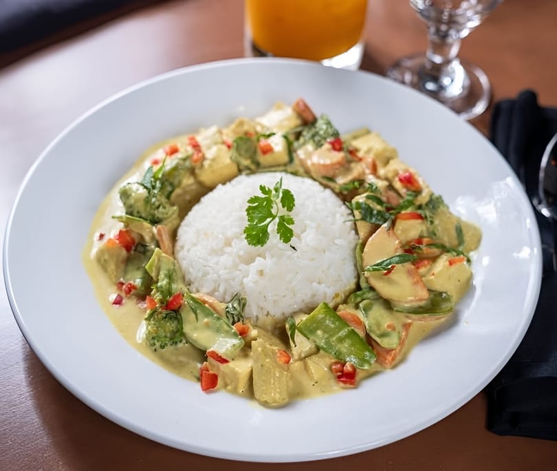 A white plate with a mound of white rice surrounded by yellow curry with vegetables and tofu.