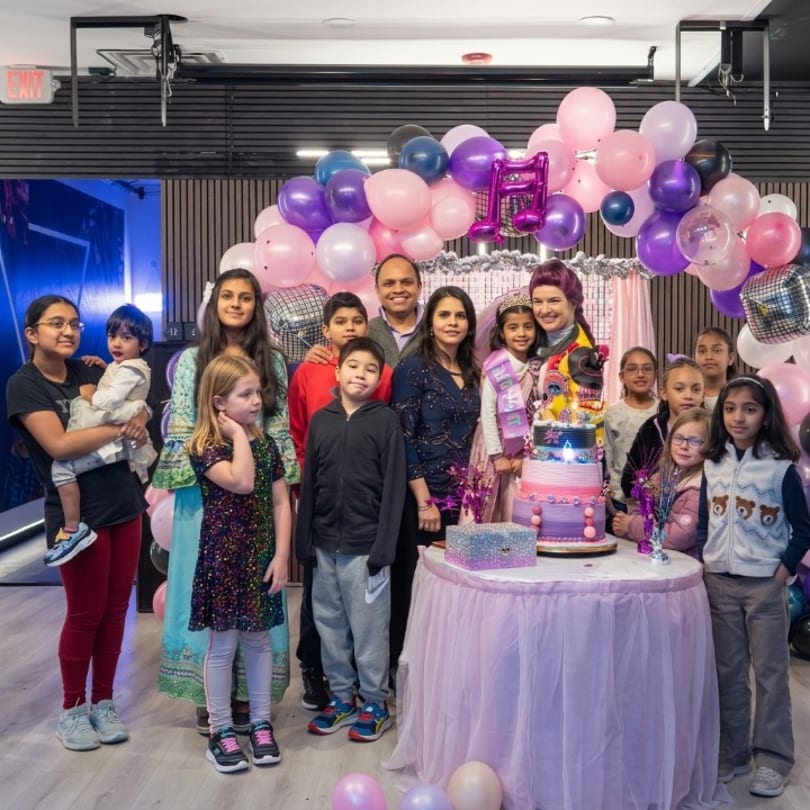Happy group of adults and children celebrating a birthday with a tiered cake and colorful balloons.