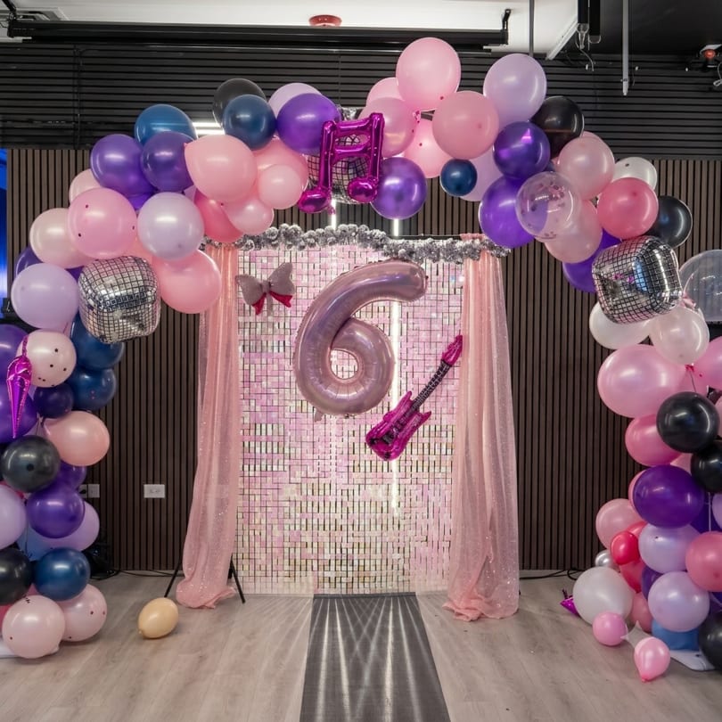 Pink, purple, and black balloon arch with disco balls frames a number 6 balloon and guitar against a sequin backdrop.