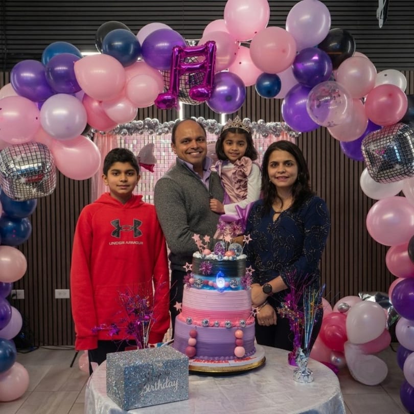 A smiling family of four stands in front of a multi-tiered birthday cake and a pink and purple balloon arch.