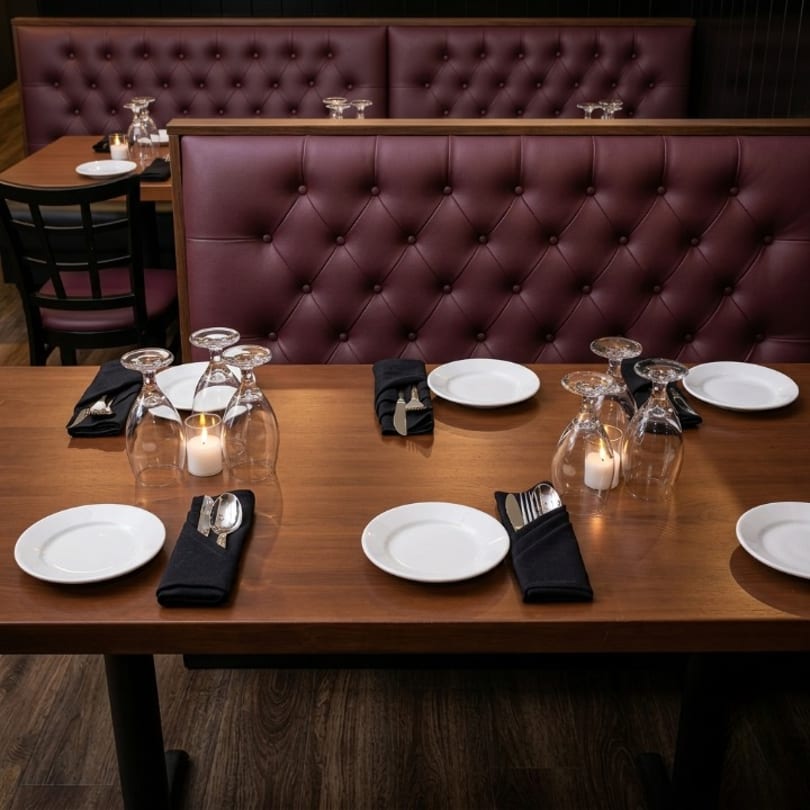 Set restaurant tables with white plates, wine glasses, candles, and elegant burgundy tufted booths.