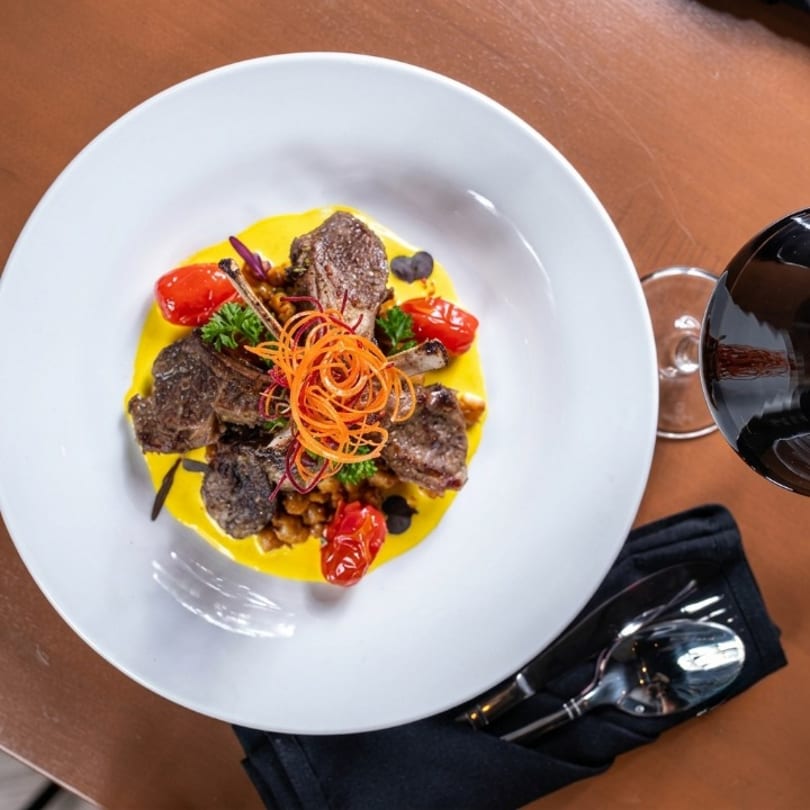 Gourmet lamb chops on yellow puree with cherry tomatoes, shredded carrots, and a wine glass.